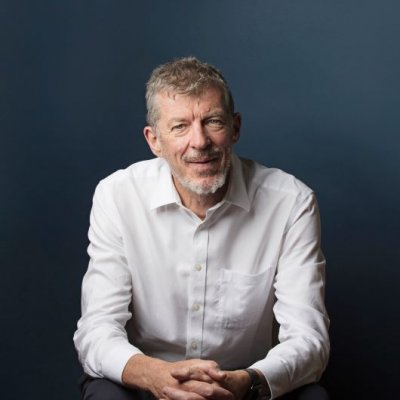 A man with grey hair and wearing a white shirt sits clasping his hands